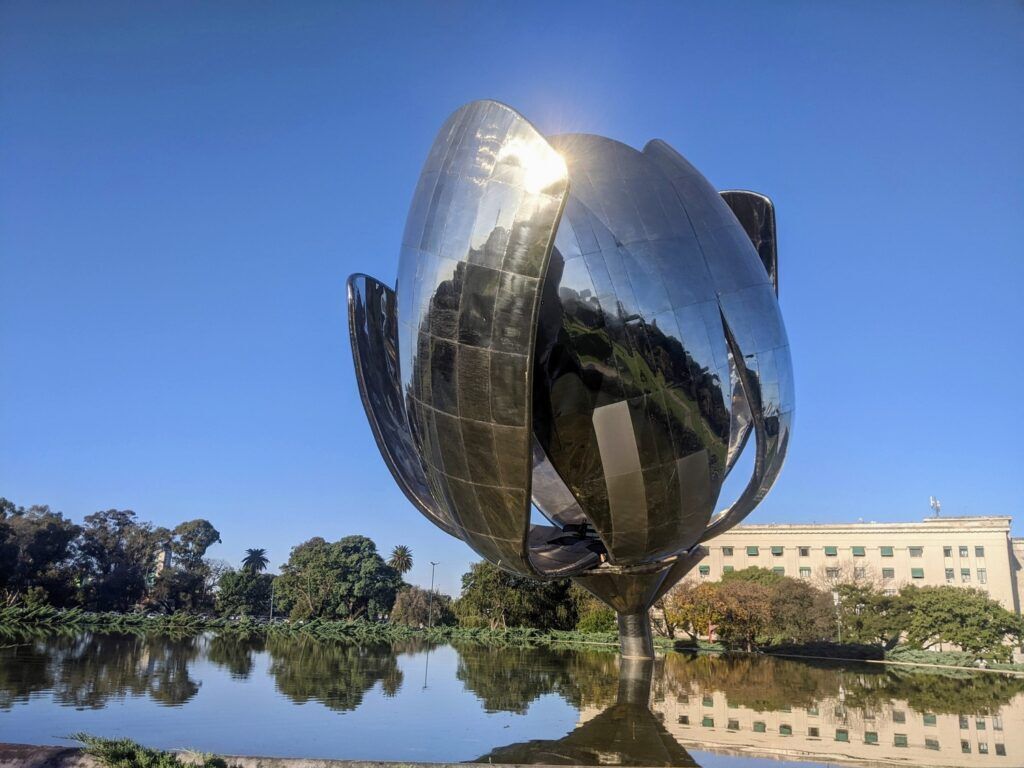 A large, reflective metal flower sculpture in an open park setting, partially opened with petals stretched outward, surrounded by grass and trees.