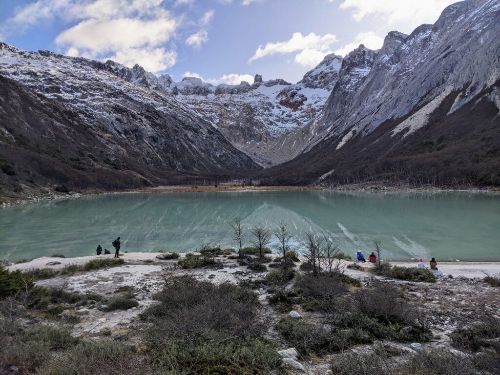 A serene mountain landscape featuring a picturesque green lagoon, surrounded by lush vegetation and rugged terrain, indicating a hiking trail location with scenic vistas.