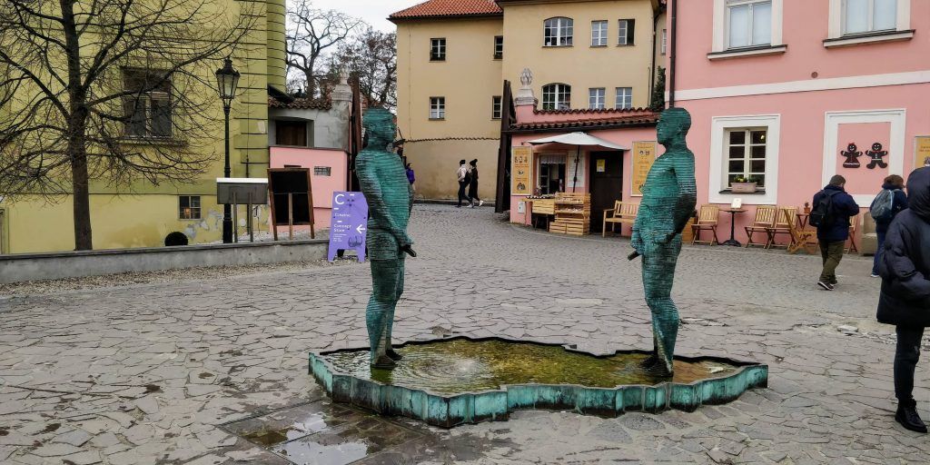 Bronze statue of a urinating man, located outdoors on a circular platform with water. The statue features a motion capturing a mid-action pose in an urban setting.