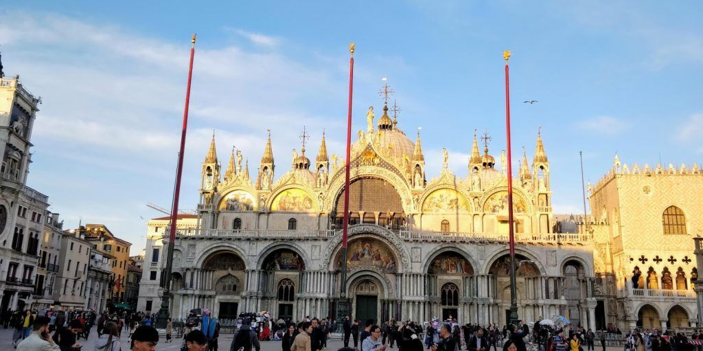 A square filled with numerous flying pigeons in a public area where people can feed them by hand, with notable architecture visible in the background, such as a basilica.