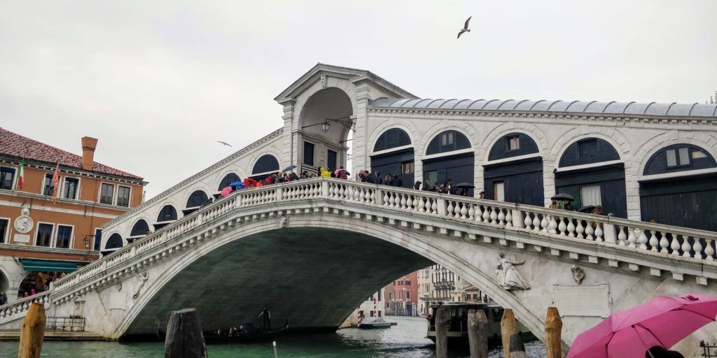 A busy street scene in Venice, with people walking and some boats visible on the water beside the sidewalk. The background includes distinctive Venetian architecture featuring arched windows and decorative details.