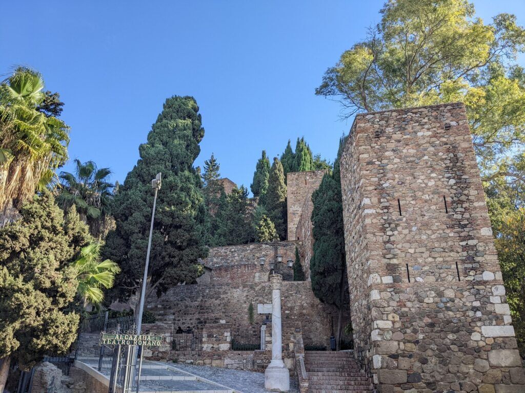 View of Malaga's Roman Theatre, an ancient stone amphitheater with seating steps, surrounded by modern city buildings and lush greenery.