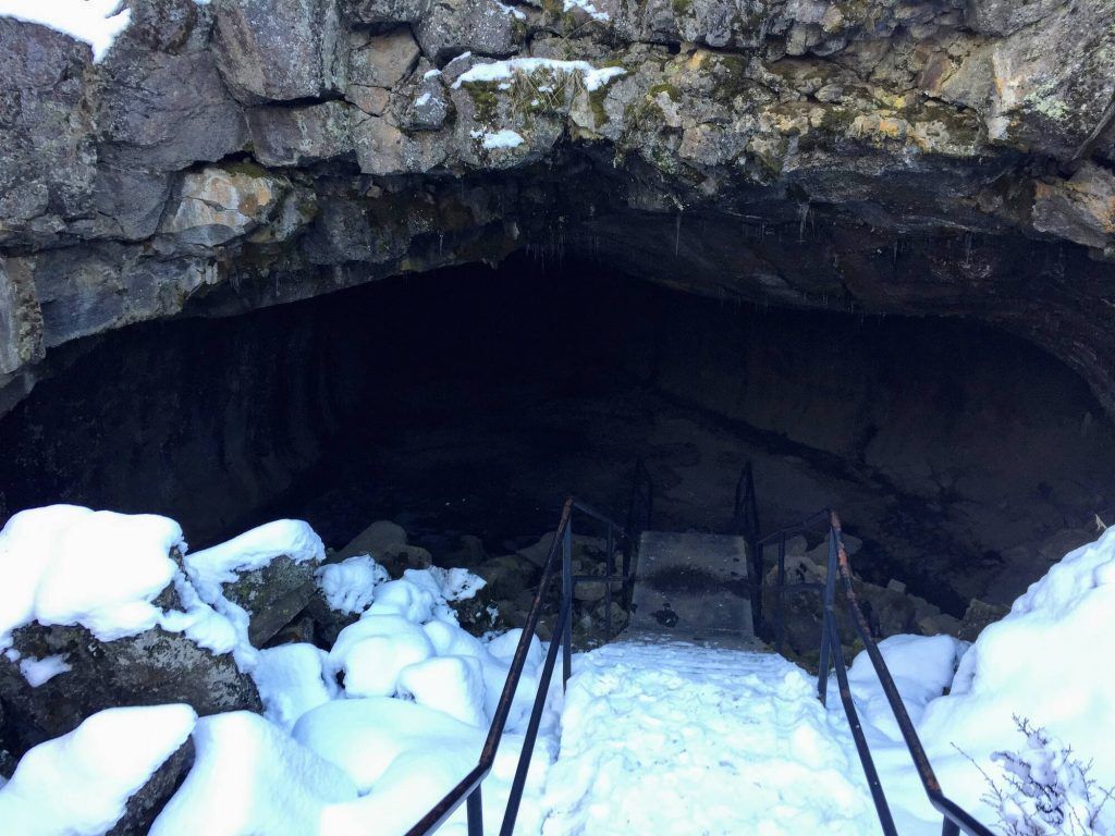 A narrow, dimly lit underground tunnel with rugged rock walls, part of a 1/3 mile easy hike. The location is associated with the Lassen Volcanic National Park.