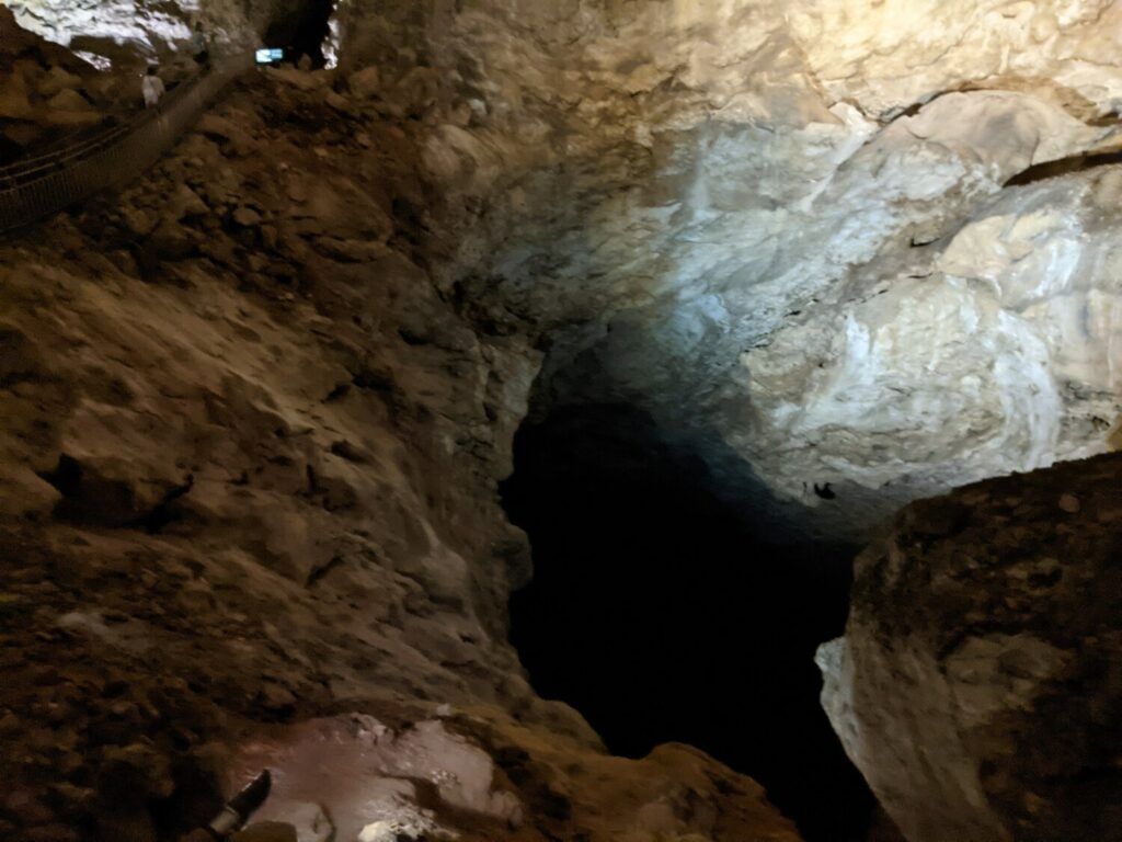A cave interior with stalactites and stalagmites illuminated by artificial lighting, creating a dramatic and textured underground landscape.